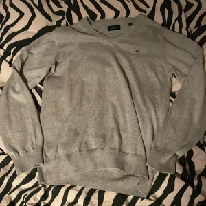 Grå tröja från Gant - Säljer en stilren grå tröja från Gant med v-ringning och broderad logga på bröstet. Tröjan har långa ärmar och ribbade muddar vid ärmslut och nederkant. Perfekt för en avslappnad look.