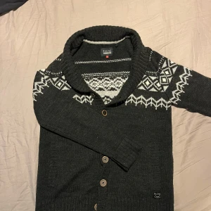 Mörkgrå stickad kofta med mönster - Säljer en mysig mörkgrå stickad kofta med ett stilrent vitt mönster över bröstet och axlarna. Koftan har knappar framtill och en bred krage som ger extra värme. Perfekt för kyliga dagar.