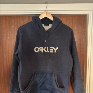 Mörkblå hoodie från Oakley - Säljer en snygg vintage mörkblå hoodie från Oakley med stor logga på bröstet. Tröjan har en klassisk känguruficka och justerbar huva med dragsko. 