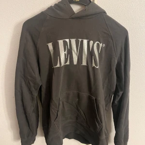 Svart hoodie från Levi's - Säljer en svart hoodie från Levi's med stor logga på framsidan. Tröjan har en klassisk design med huva och en stor ficka framtill. Perfekt för en avslappnad stil. Tyvärr så saknar den snöret till huvan men annars i fint skick:)