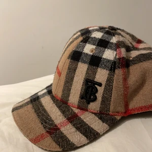 Rutig keps från Burberry - Snygg rutig keps från Burberry i beige, svart och röd. Kepsen har ett broderat Burberry-märke på baksidan och är justerbart där bak. Perfekt för att ge din outfit en stilren touch.