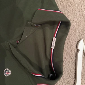 Grön pikétröja från Moncler - Snygg grön pikétröja från Moncler med klassisk krage och knappar. Tröjan har röda och vita detaljer på kragen och ärmsluten samt Moncler-logga på bröstet. Perfekt för en stilren look. Liten i storlek, mer åt L/M hållet