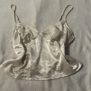 Vit satinlinne från H&M Divided - Säljer ett elegant vitt satinlinne från H&M Divided. Linnet har smala axelband och spetsdetaljer vid bysten samt en liten rosett framtill. Perfekt för en stilren look.