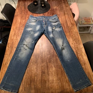 Blå jeans från Dsquared2 - Säljer ett par blå jeans från Dsquared2 med coola slitningar och en unik 'Eviction Notice'-lapp på baksidan. Jeansen har en straight passform och klassisk femficksdesign. Perfekta för dig som vill ha en edgy look! 