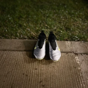 Nike fotbollsskor i silver och neon - Snygga Nike fotbollsskor i silver med neonfärgade detaljer och svart Nike-logga. Skorna har en modern design med textmönster och är perfekta för fotbollsplanen. De har en tight passform och dobbar för bra grepp.