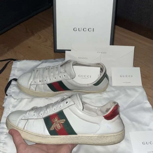 Vita sneakers från Gucci - Säljer ett par vita sneakers från Gucci med klassiska gröna och röda ränder samt en broderad guldfärgad insekt på sidan. Skorna har snörning och en låg profil, perfekt för en stilren look. Obs! Allt ingår, extra laces, kort och box inte dustbag