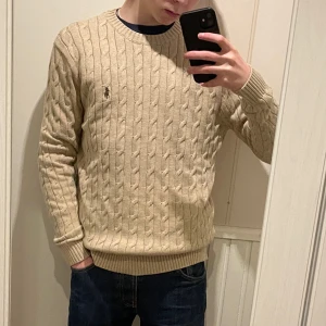 Beige stickad tröja från Ralph Lauren - Säljer en stilren beige kabelstickad tröja från Ralph Lauren. Tröjan har en klassisk rund halsringning och långa ärmar med ribbade muddar. Perfekt för en avslappnad och snygg look. Lite större i storleken, han på bilden är 175cm. 💰 (Pris går att diskuteras vid snabb affär)💰 INGA RETURER!🚨