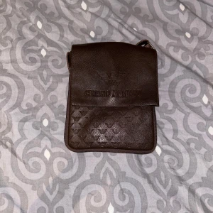 Brun axelväska från Giorgio Armani - Snygg brun axelväska från Giorgio Armani i skinn. Väskan har ett diskret mönster och en praktisk design med lock. Perfekt för att bära med sig det nödvändigaste med stil.