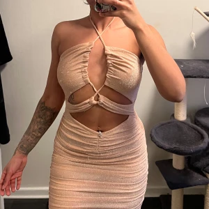 Glittrig klänning - Snygg glittrig beige/rosé klänning med halterneck och cut-out detaljer. Klänningen har en tight passform och är kort, perfekt för en festlig kväll. Den har en knytning i ryggen och en smickrande rynkad design.
