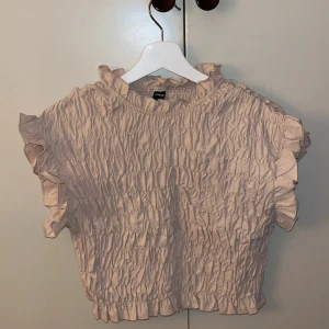 Beige blus från Shein - Säljer en söt beige blus från Shein med volangdetaljer. Blusen har en rynkad design och korta ärmar med volanger vid ärmslut och hals. Perfekt för en stilren look.