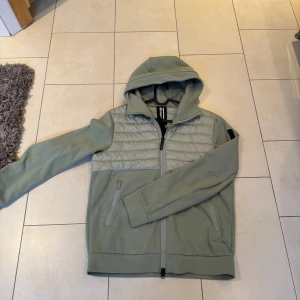 Grön cardigan från Bomboogie - Snygg grön cardigan  från Bomboogie med huva och dragkedja. Cardiganen har quiltade detaljer på bröstet och är tillverkad i syntetmaterial. Perfekt för en stilren look.