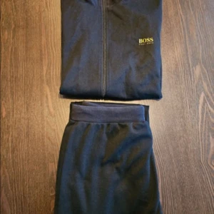 Svart träningsset från Hugo Boss - Snyggt svart träningsset från Hugo Boss med dragkedja på jackan och matchande byxor. Jackan har långa ärmar och praktiska fickor med dragkedja. Perfekt för en stilren look under träningen eller en avslappnad dag. Står s men passar som m/l