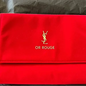 Röd kuvertväska från Yves Saint Laurent - Snygg röd kuvertväska från Yves Saint Laurent med guldlogga på framsidan. Väskan har en elegant design med en knappstängning och svart insida. Perfekt för att ge en touch av lyx till din outfit.