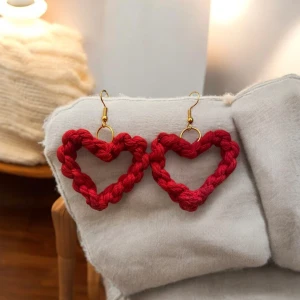 Röda hjärtformade örhängen - Charmiga röda örhängen i form av hjärtan. De är knutna med makramé och hänger från en guldfärgad krok. Perfekta för att ge en romantisk touch till din outfit.