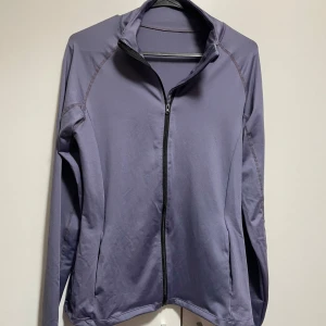 Lila träningströja med dragkedja - Snygg lila träningsjacka med dragkedja framtill. Jackan har en hög krage och långa ärmar, perfekt för träning eller en aktiv vardag. Den är tillverkad i ett syntetmaterial som andas och ger bra rörelsefrihet. Jätte bekväm till gymmet!