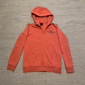 Orange hoodie från Peak Performance - Säljer en snygg orange hoodie från Peak Performance med dragkedja och huva. Den har en broderad logga på bröstet och är tillverkad i ett mjukt material. Perfekt för en avslappnad stil.