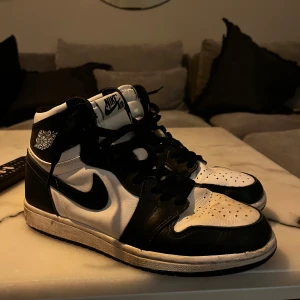 Jordan 1 - Snygga Nike Air Jordans i klassisk svart och vit färgkombination. Skorna har en hög modell med snörning och den ikoniska Nike-loggan på sidan.