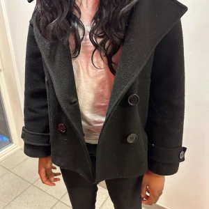 Svart kappa med knappar - Snygg svart kappa med stora knappar och bred krage. Perfekt för kyligare dagar. Den har långa ärmar och en klassisk design som funkar till det mesta. Ett måste i garderoben för den stilmedvetna.