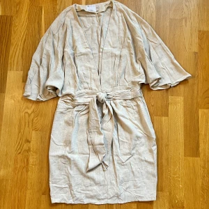 Linneklänning med knytband - Säljer en stilren beige klänning med knytband i midjan. Klänningen har en v-ringad design och vida ärmar som ger en luftig känsla. Perfekt för en avslappnad och chic look. 