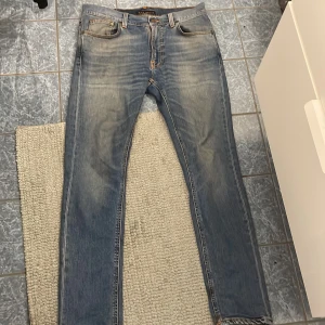 Blå jeans från Nudie Jeans - Säljer ett par klassiska blå jeans från Nudie Jeans. De har en snygg tvätt med lätt slitna detaljer och orange sömmar. Perfekt för en avslappnad stil. Midjan är normal och passformen är lite slim. Har andvänds 1 gång