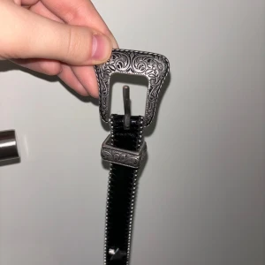 Svart bälte med dekorerat spänne från Topshop - Snyggt svart bälte från Topshop med ett detaljerat, silverfärgat spänne. Bältet har en rad med små nitar längs kanterna som ger det en extra touch. Perfekt för att ge din outfit lite edge.