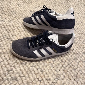 Adidas Gazelle Mörkblå mocka - Snygga Adidas Gazelle sneakers i mörkblå mocka.   Box och ett par vita snören tillkommer. Mockan är inte helt frisk men går nog att fixa med mocka borste(inte garanterat dock) Sulan är använd som du kan se på bilden. Men annars är det en ren sko med vita sulor och utan fläckar.