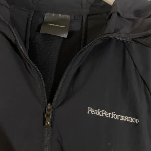Svart vindjacka från Peak Performance - Säljer en svart vindjacka från Peak Performance med dragkedja och huva. Jackan har långa ärmar med reflekterande detaljer och en praktisk ficka med dragkedja på sidan. Perfekt för blåsiga dagar.