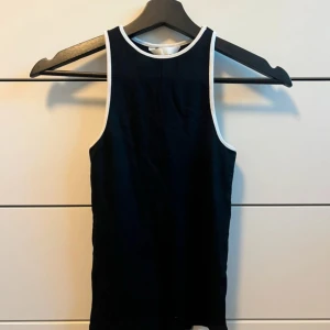 Svart ärmlös linnetopp med vita kanter - Snygg svart ärmlös linnetopp med kontrasterande vita kanter runt ärmhål och halsringning. Perfekt för varma dagar eller som ett lager under en jacka.