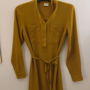 Gul klänning med knytband - Snygg gul klänning med knytband i midjan och knappar framtill. Den har långa ärmar och två bröstfickor som ger en stilren look. Perfekt för en färgglad touch i garderoben! Går ner till knäna. Perfekt för en utekväll på sommaren 