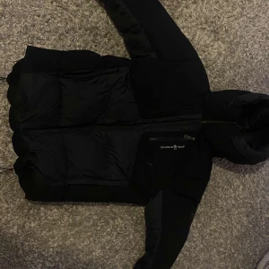 Sail racing jacka S - Snygg svart dunjacka från Sail Racing med dragkedja och huva. Perfekt för kyliga dagar med sin quiltade design som håller värmen. Jackan har en stilren look och är ett måste för garderoben.