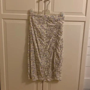 Blommig kjol med snörning - Charmig blommig kjol i ljusa toner av lila och gul med snörningsdetalj på sidan. Kjolen har en avslappnad passform och är perfekt för varmare dagar. Storleken är M, men passar som S. Från Hollister. I fint skick. 