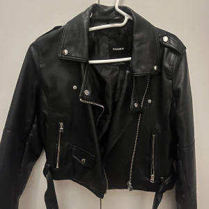 Svart skinnjacka från Pull&Bear - Snygg svart skinnjacka från Pull&Bear med coola dragkedjedetaljer och knappar. Jackan har en klassisk bikerstil med bälte i midjan och axelklaffar. Perfekt för att ge din outfit en edgy touch.