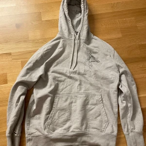 Grå hoodie från Palace - Säljer en grå hoodie från Palace med tryck på både fram- och baksidan. Hoodien har en klassisk känguruficka och justerbar huva med dragsko. Perfekt för en avslappnad stil. Trasig i ärmen samt ett litet hål vid fickan.
