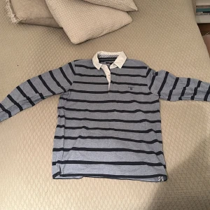 Blå och grå randig Gant pikétröja - Säljer en stilren blå och grå randig pikétröja med krage och knappar. Perfekt för en avslappnad men ändå snygg look. Tröjan har långa ärmar och är gjord i ett mjukt material.