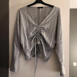 Grå ribbad tröja med knyt - Säljer en snygg grå ribbad tröja med v-ringning och knyt framtill. Tröjan har långa ärmar och en trendig design som passar perfekt för kyligare dagar.