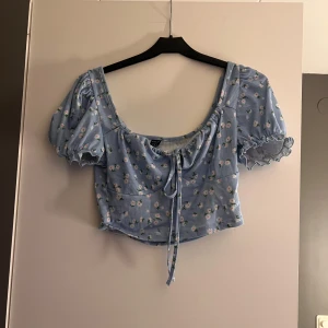 Blå blommig croppad blus - Söt blå blommig blus från Bershka med puffiga korta ärmar och knytning framtill. Perfekt för en somrig look med sitt lätta och luftiga material.