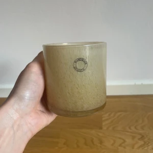 Beige doftljus i glas - Säljer ett stilrent ljushållare i glas med en rund etikett på framsidan. Perfekt för att skapa en mysig atmosfär hemma. Glaset har en mjuk, naturlig ton som passar in i de flesta inredningar.