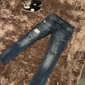 Blå jeans från Dsquared2 - Snygga blå jeans från Dsquared2 med slitna detaljer och färgstänk. De har en klassisk femficksdesign och knappgylf. Perfekta för en avslappnad stil med en touch av edge. Slim fit ny pris 7000 köpta på Vestiaire Collective st 30:32