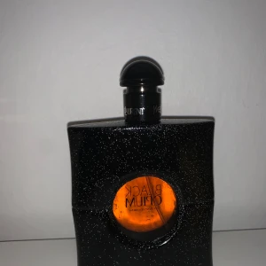 Black Opium parfym från Yves Saint Laurent - Snygg och elegant parfymflaska i svart med glittrande yta och en rund, orange detalj i mitten. Flaskan har en unik form och en svart kork. Perfekt för den som älskar en djärv och mystisk doft.