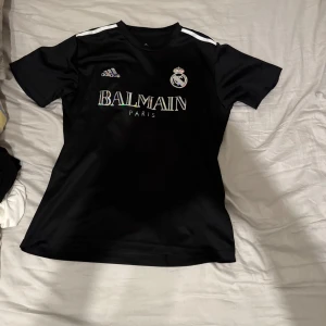 Balmain X Real Madrid  - Snygg svart fotbollströja från Adidas med vit logga och texten 'Balmain Paris' på framsidan. Tröjan har korta ärmar och tre vita ränder på axlarna. Perfekt för fotbollsfans som vill ha stil på planen. Pris kan diskuteras
