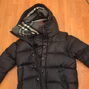 Svart Burberry dunjacka - Säljer en stilren svart dunjacka från Burberry med klassiskt rutigt foder. Jackan har en hög krage och stängs med både dragkedja och knappar. Perfekt för kyliga dagar.