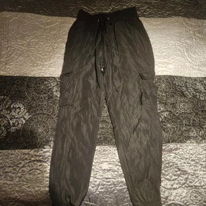 Svarta cargobyxor - Säljer ett par svarta cargobyxor med elastisk midja och snörning. Byxorna har flera praktiska fickor och resår vid bensluten för en bekväm passform. Perfekta för en avslappnad stil.