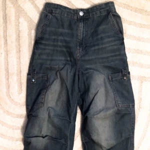 Blå cargopants med fickor - Säljer ett par blå cargopants med flera praktiska fickor och elastisk midja. Byxorna har en loose passform och är perfekta för en avslappnad stil. De är tillverkade i jeansmaterial och har en klassisk design med knapp och dragkedja framtill.