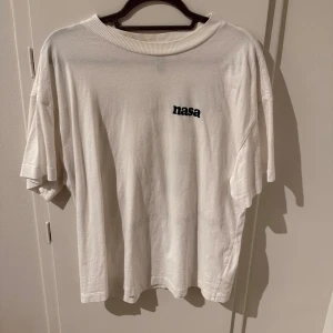 Vit NASA t-shirt - Säljer en vit t-shirt med NASA-tryck. T-shirten har en normal passform och korta ärmar. På framsidan finns en liten NASA-logga och på baksidan ett större tryck med texten 'Comet Cluster' och 'Deep Space Orbit'. Perfekt för rymdentusiaster!