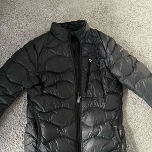 Svart dunjacka från Peak Performance - Snygg svart dunjacka från Peak Performance med dragkedja och praktiska fickor. Perfekt för kyliga dagar med sin quiltade design som håller värmen. Jackan har en stilren look och är lätt att matcha med olika outfits. 