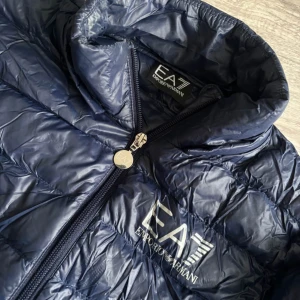 Mörkblå pufferjacka från Emporio Armani - Storleken är lite oklar, men jag tippar att man kan ha den om man har 34-36. Den är figursydd. Snygg mörkblå pufferjacka från Emporio Armani med dragkedja och hög krage. Perfekt för kyliga dagar med sin quiltade design och stilrena look. 