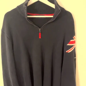 Marinblå tröja med dragkedja och flaggdetalj - Snygg svart tröja med dragkedja och ribbad krage. Har en cool flaggdetalj på ärmen i rött och vitt. Perfekt för en stilren och avslappnad look.