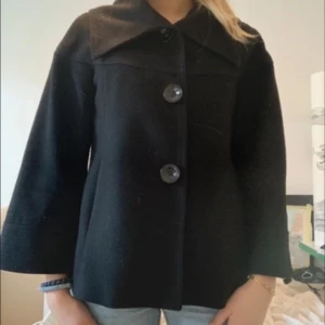 Svart kappa med stora knappar - Snygg svart kappa med stora knappar framtill. Den har en klassisk krage och trekvartsärmar, vilket ger en stilren look. Perfekt för höst och vår.
