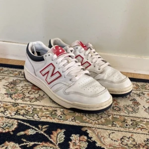 Vita sneakers från New Balance och de har svarts skosnören nu - Säljer ett par vita New Balance sneakers med röda och blå detaljer. Skorna har snörning och en bekväm sula, perfekt för vardagsbruk. Den klassiska New Balance-loggan syns tydligt på sidan.
