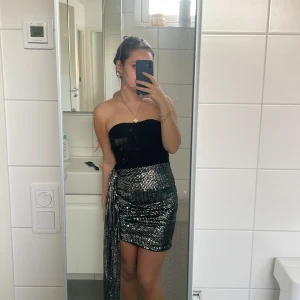 Svart och silver axelbandslös klänning - Trendig axelbandslös klänning i svart och silver med glittriga paljetter. Den har  slits  på sidan som hänger ner. Perfekt för nyår!! OBS: jag är 154 cm lång, därav är slitsen ganska lång för mig. ALDRIG använt, kände tyvärr att slitsen var ganska lång för min smak. 